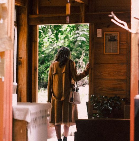 kobieta w brązowym płaszczu stojąca w otwartych drzwiach drewnianego domku patrząc na liście